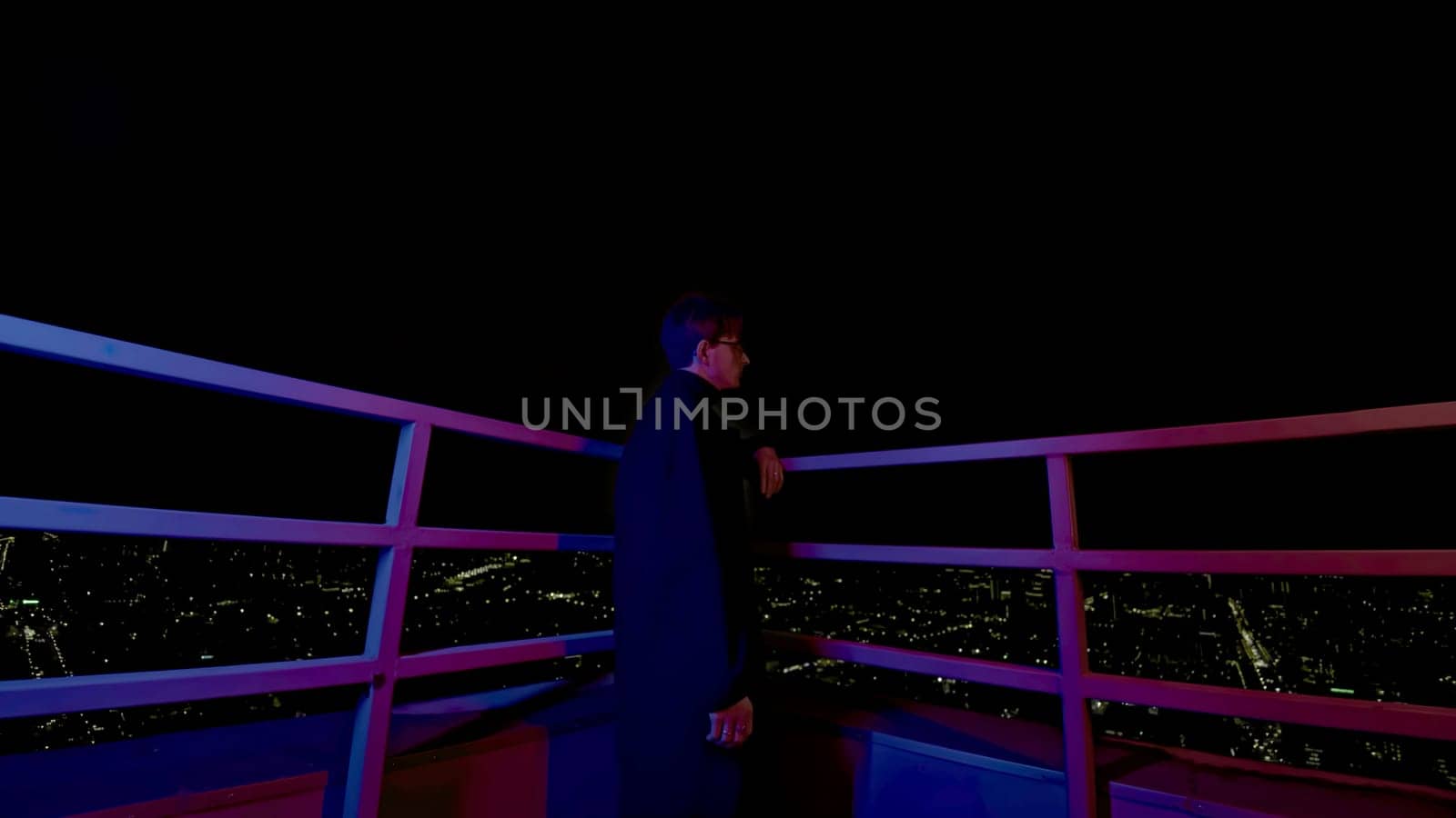 Stylish young man looks at city from roof. Media. Fashion man in coat on roof of skyscraper overlooking city. Young man looks at night city from roof of high-rise building.