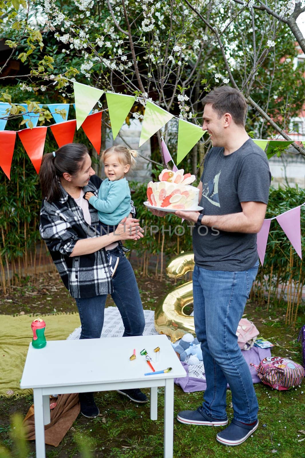 Little smiling girl sits in the arms of her mother standing next to her father with a birthday cake in his hands. High quality photo