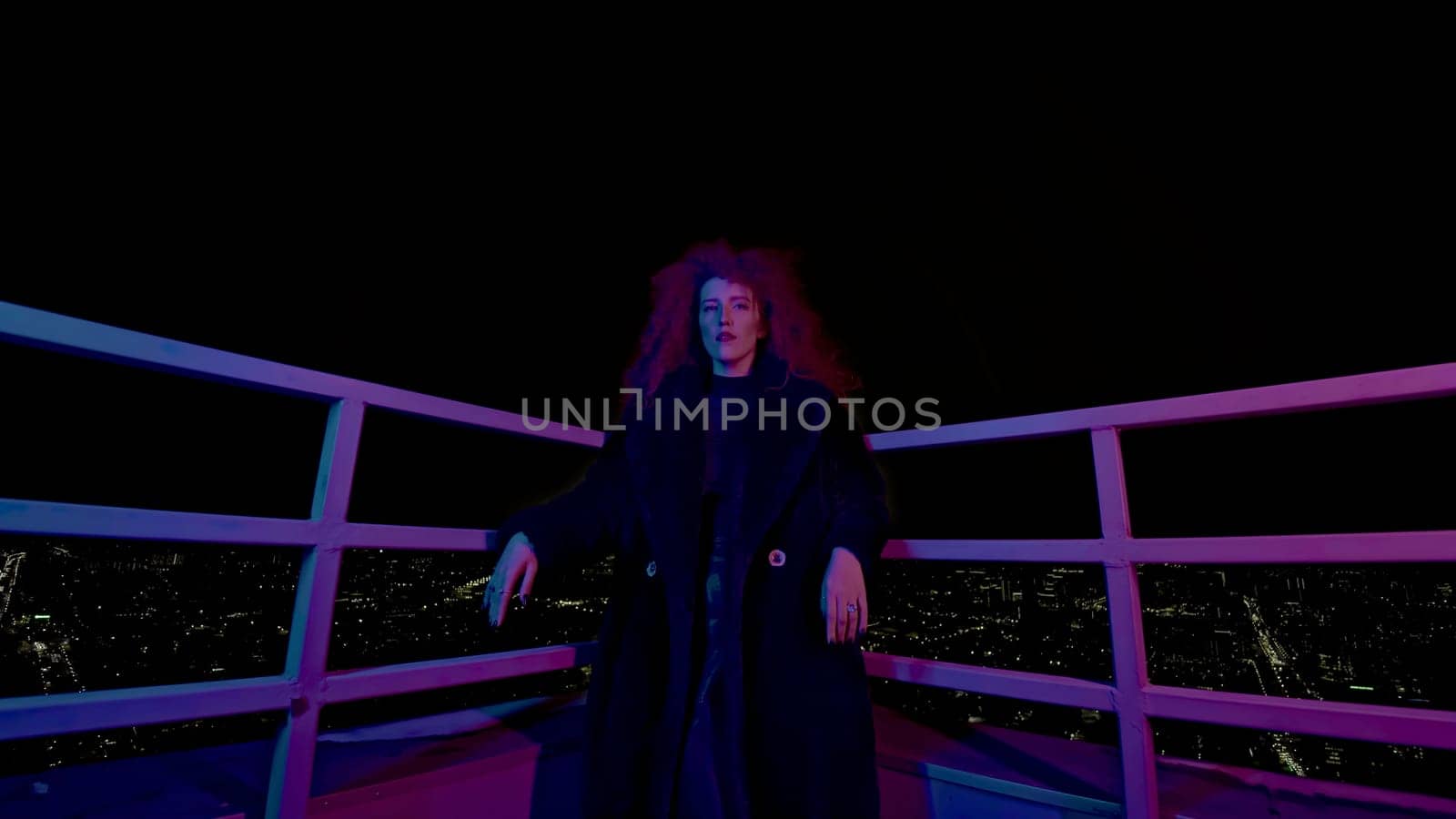 Woman on roof of night city. Media. Stylish fashion shoot with model in coat on roof. Female model in neon night shooting on roof of high-rise.