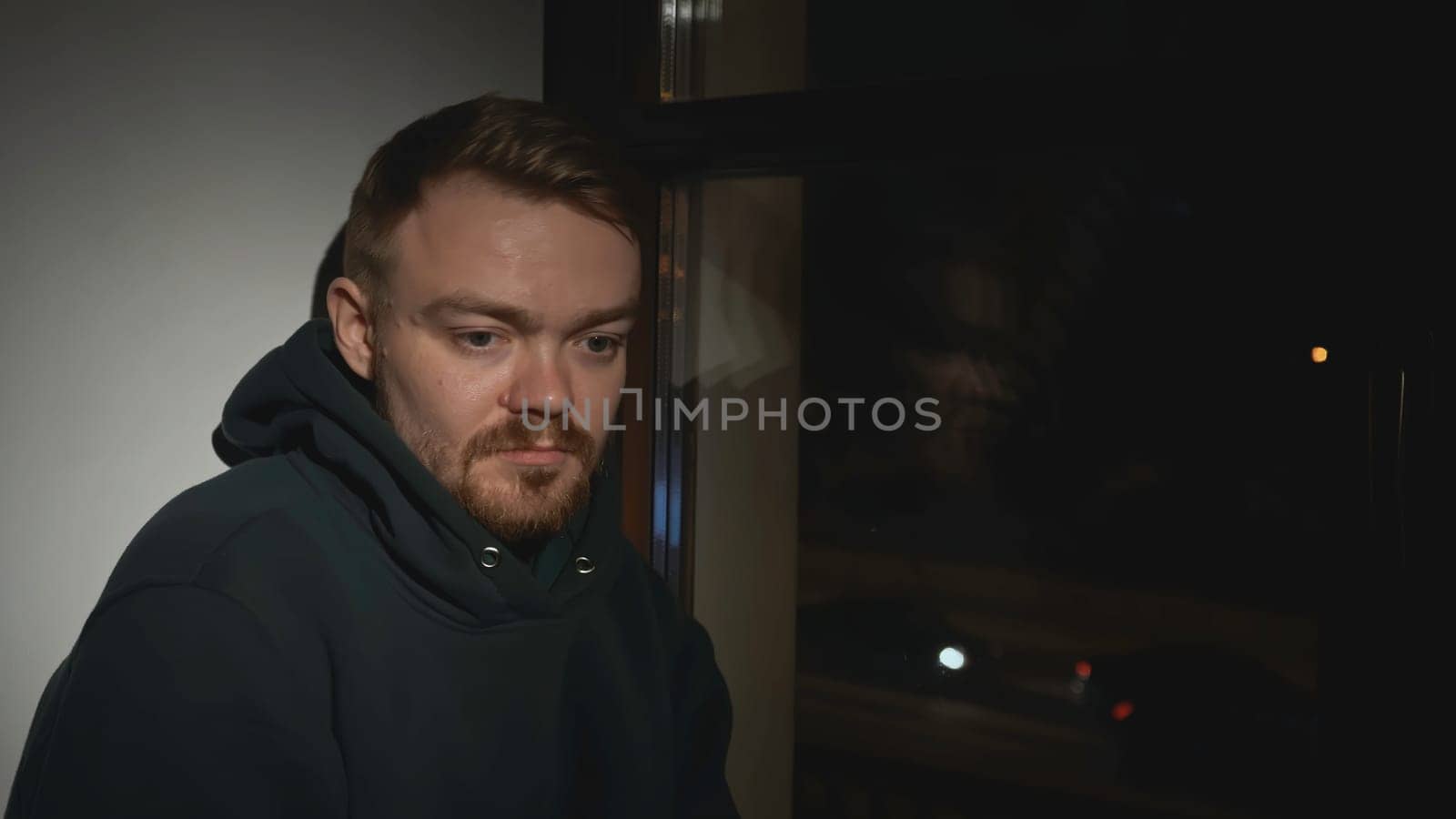 Sad man at night window of house. Media. Man looks out window with sad look and goes to sleep. Tired lonely man looks out window of house.