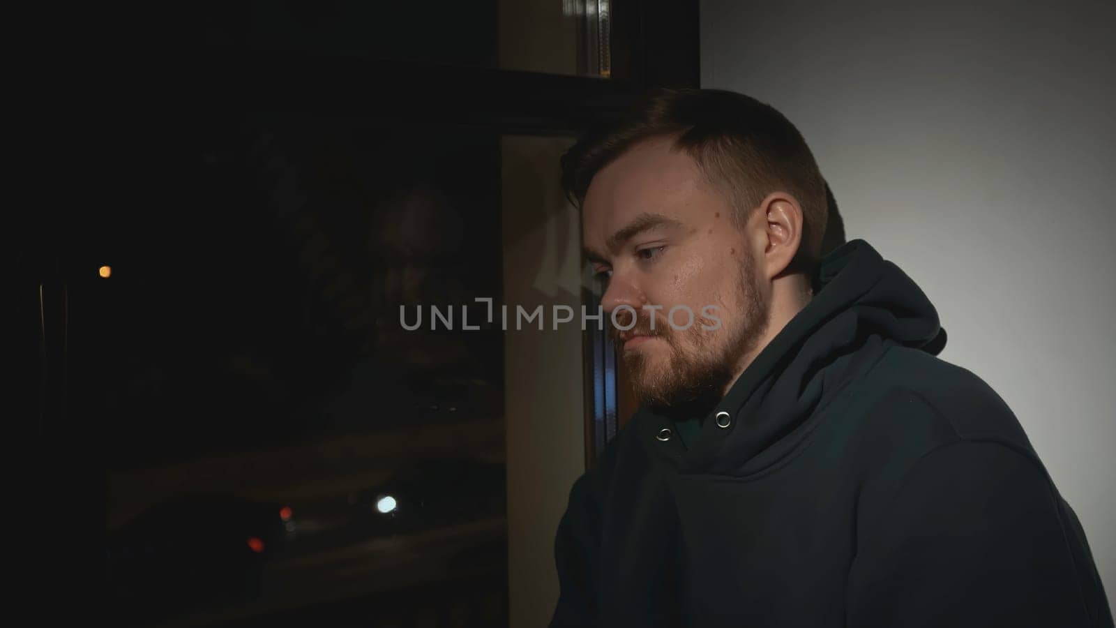 Sad man at night window of house. Media. Man looks out window with sad look and goes to sleep. Tired lonely man looks out window of house.