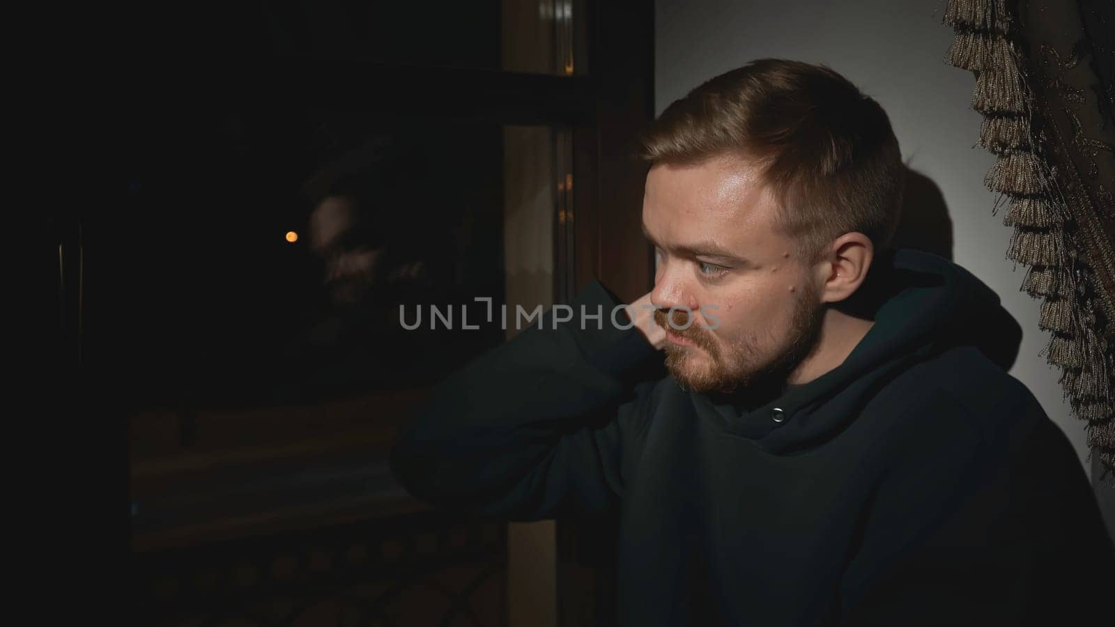 Young man sits tired and looks out window. Media. Lonely man looks out window of house and is sad. Man is nervous and sad looking out night window of house.