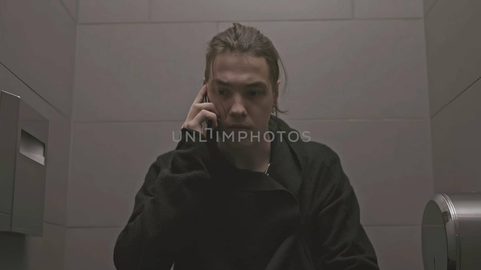 Young man sits on toilet and talks on phone. Stock. Man in black walks into toilet stall and talks on phone. Young man is talking aggressively on phone in toilet.