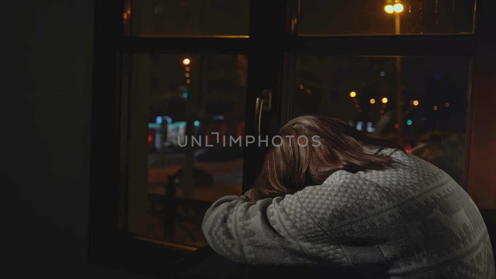 Lonely young woman looks out night window. Media. Young woman sits on window and looks sadly at night city. Woman with depression sits alone on window of house.