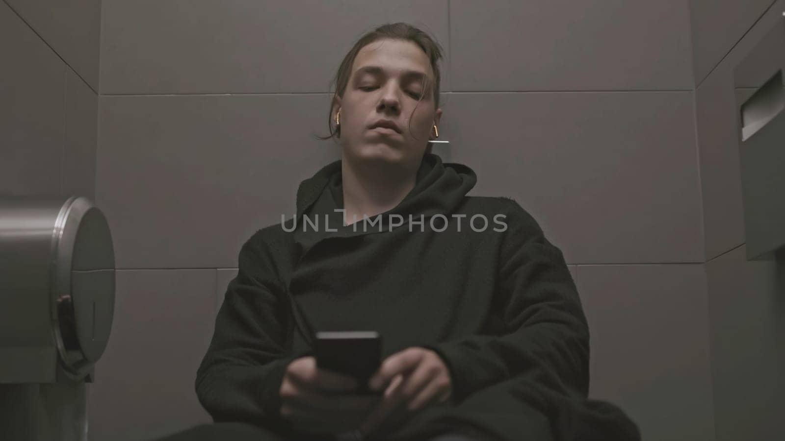 Young man with depression in toilet. Stock. Man sits on toilet and listens to sad music. Teenager hid in toilet stall and is sad.