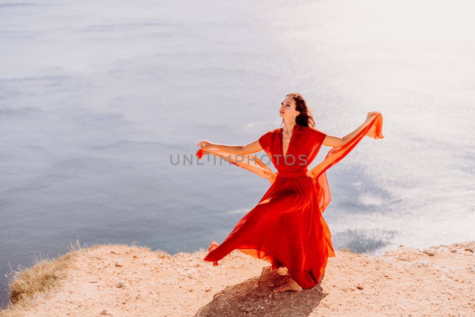 Woman red dress sea. Female dancer posing on a rocky outcrop high above the sea. Girl on the nature on blue sky background. Fashion photo