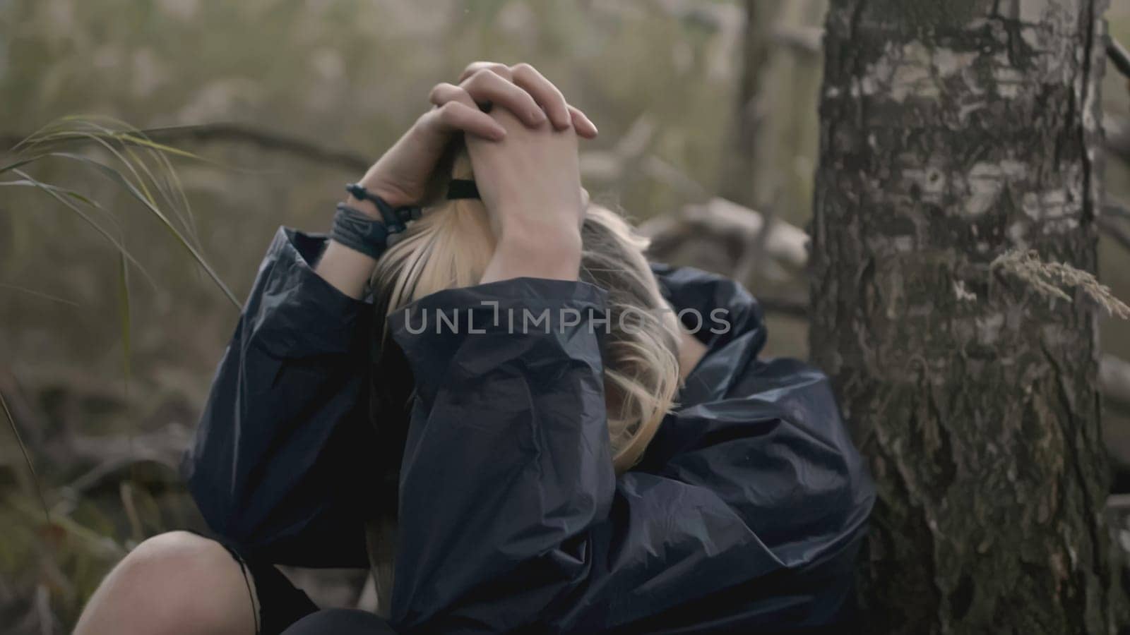 Young man sits tired by tree in forest. Stock. Young man is lost in forest and sits in despair under tree. Man is sitting by tree with desperate look and loss.