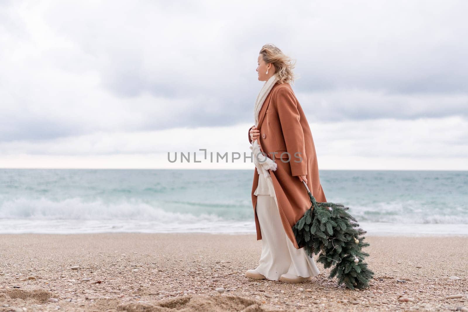 Blond woman Christmas sea. Christmas portrait of a happy woman walking along the beach and holding a Christmas tree in her hands. She is wearing a brown coat and a white suit