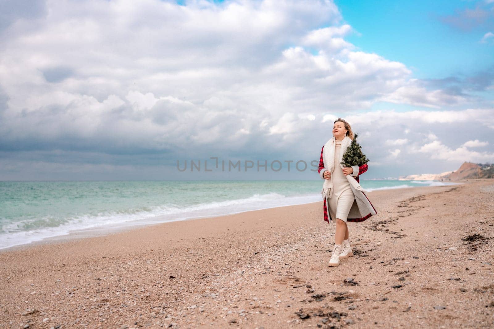 Blond woman holding Christmas tree by the sea. Christmas portrait of a happy woman walking along the beach and holding a Christmas tree in her hands. Dressed in a red coat, white dress. by Matiunina