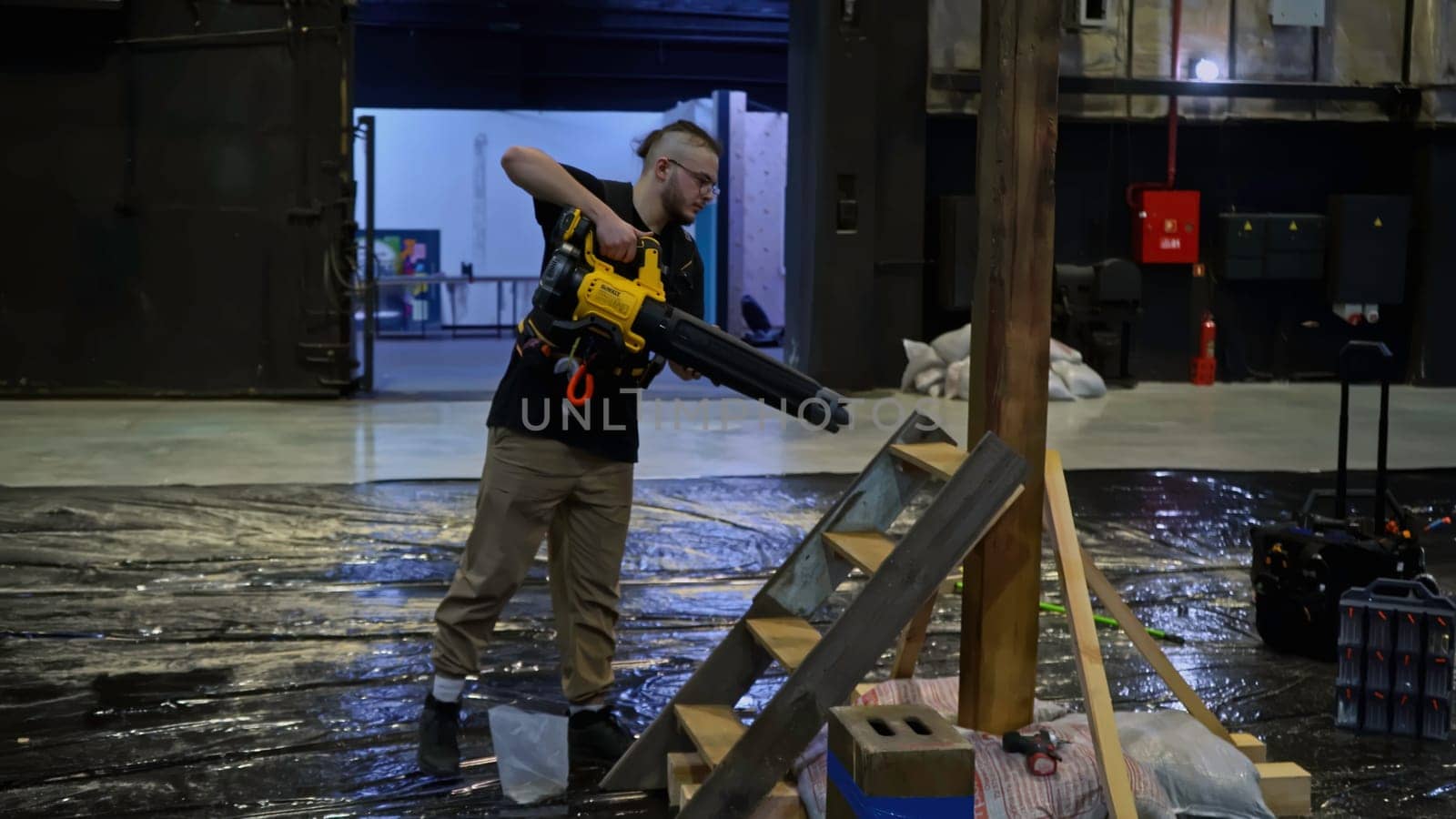 Young male worker using construction blower to clean assembled wooden stairs and pillar from sawdust, wood shavings. Media. Preparation of the stage for the concert