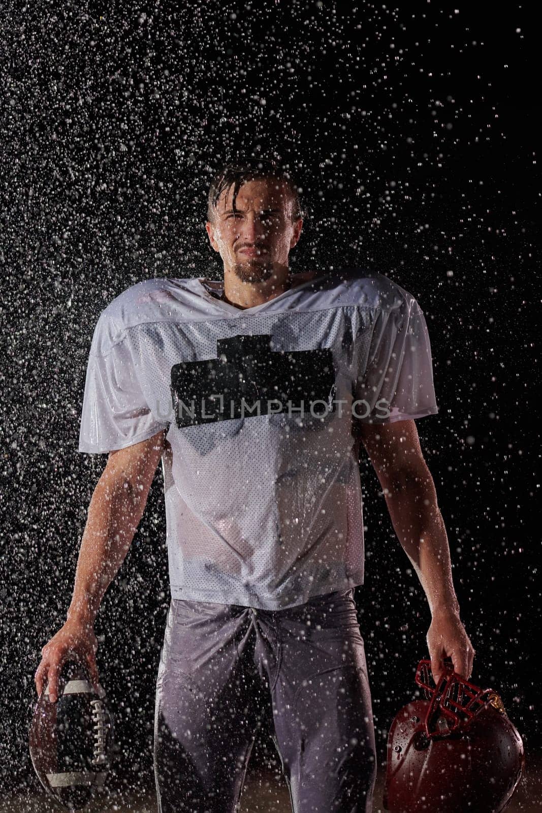 American Football Field: Lonely Athlete Warrior Standing on a Field Holds his Helmet and Ready to Play. Player Preparing to Run, Attack and Score Touchdown. Rainy Night with Dramatic Fog, Blue Light by dotshock