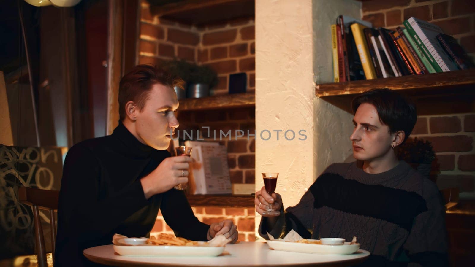 Two friends are sitting in restaurant, chatting and drinking alcoholic drink. Media. Two young men are drinking alcoholic tincture in restaurant. Cozy meeting of two friends in restaurant with alcoholic drink.