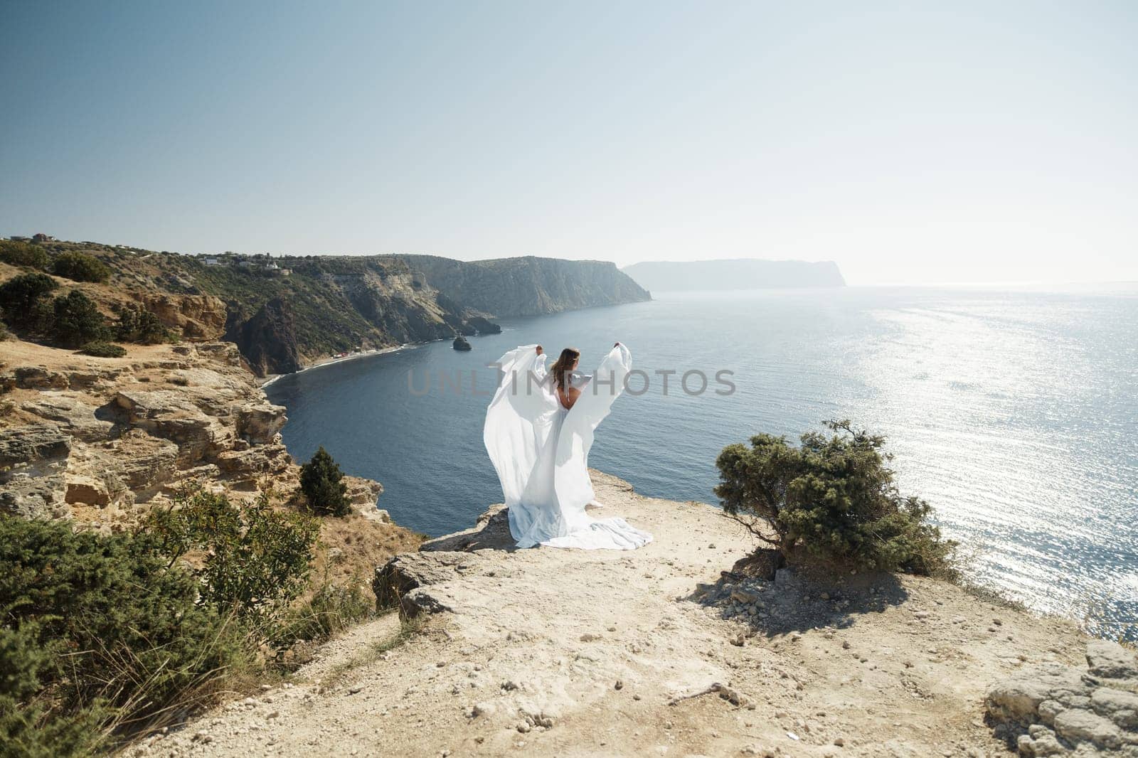 woman white dress stands on a rocky cliff overlooking the ocean. The scene is serene and peaceful, with the woman's dress billowing in the wind. The ocean in the background is calm and blue