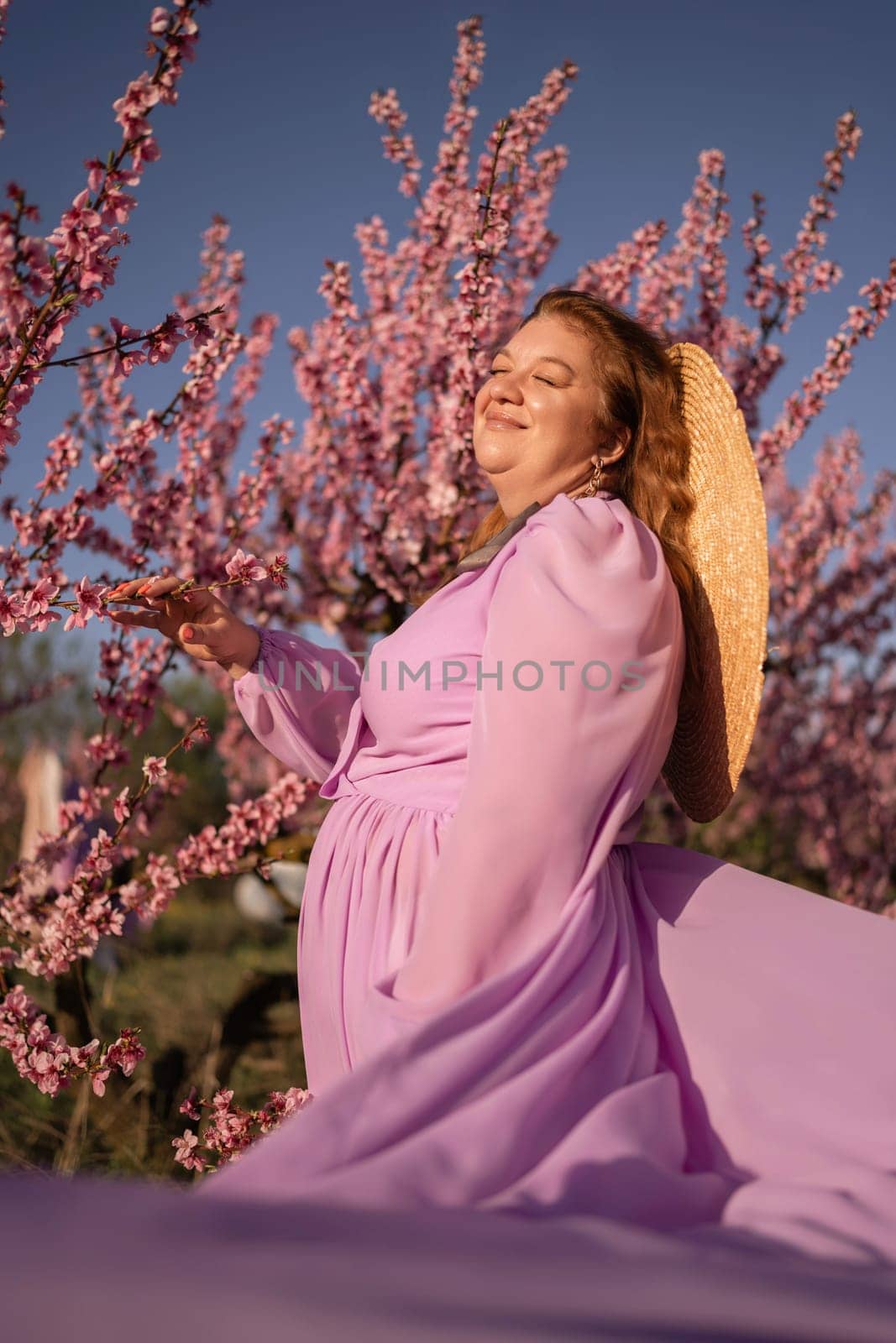 Woman blooming peach orchard. Against the backdrop of a picturesque peach orchard, a woman in a long pink dress and hat enjoys a peaceful walk in the park, surrounded by the beauty of nature