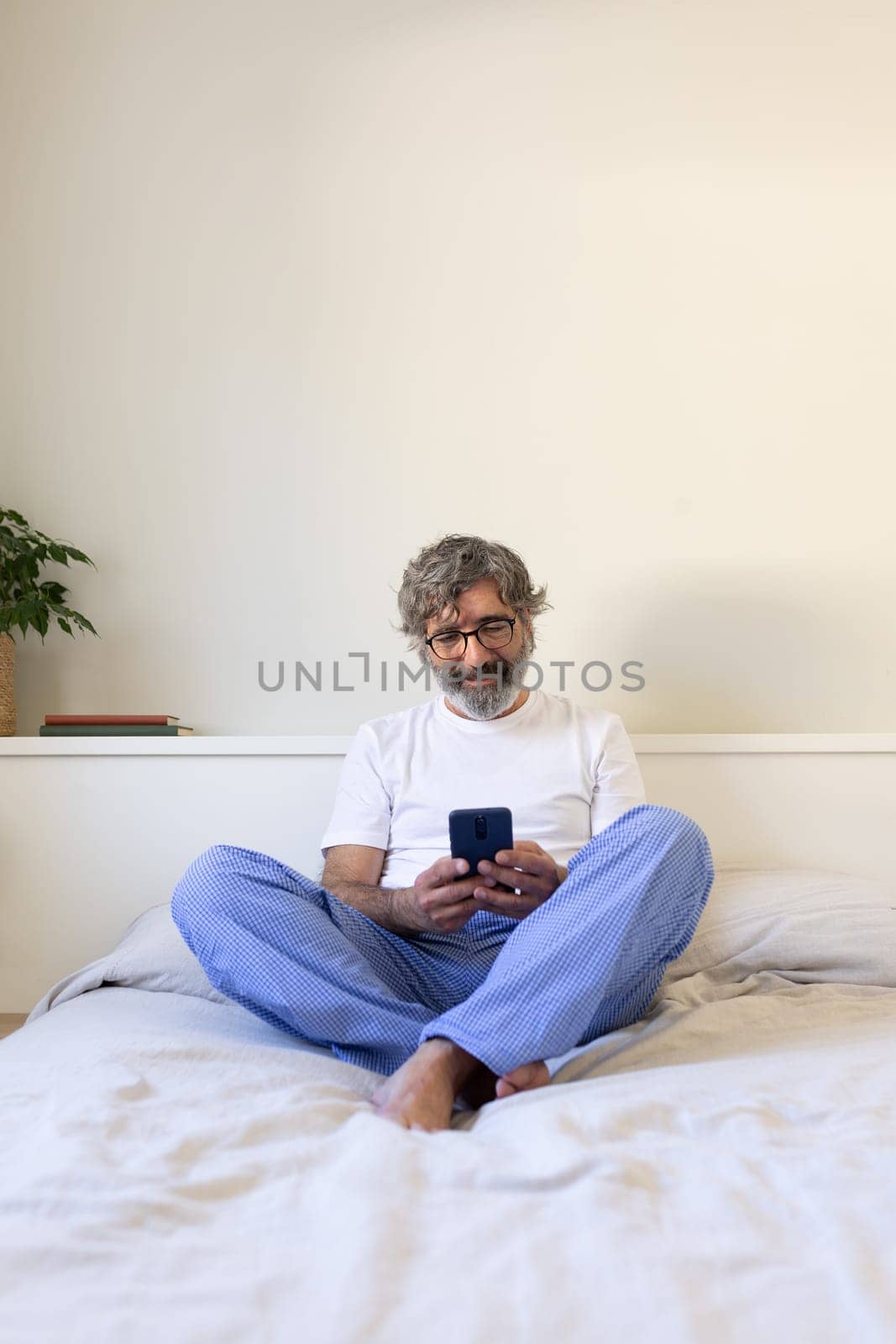 Vertical portrait of mature man using phone relaxing on bed at home. Copy space. by Hoverstock