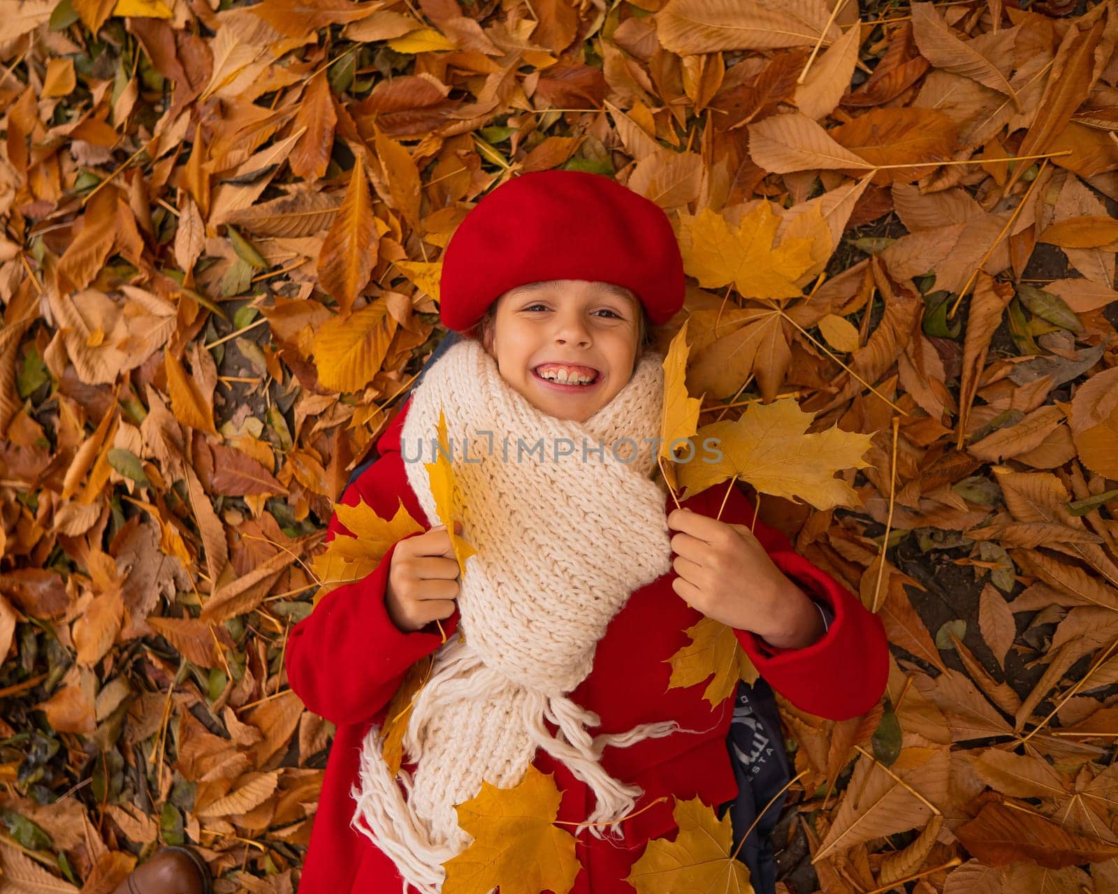 An overhead view of a caucasian girl in a red coat and beret lies on yellow foliage. Walk in the park in autumn