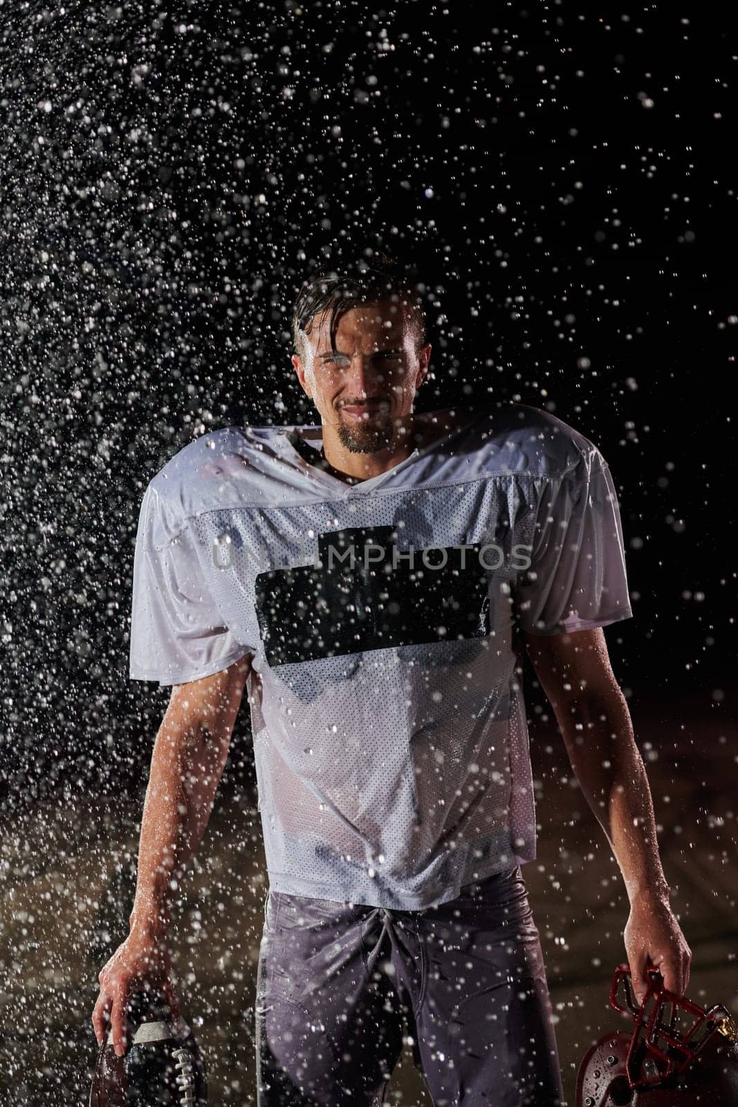 American Football Field: Lonely Athlete Warrior Standing on a Field Holds his Helmet and Ready to Play. Player Preparing to Run, Attack and Score Touchdown. Rainy Night with Dramatic Fog, Blue Light by dotshock