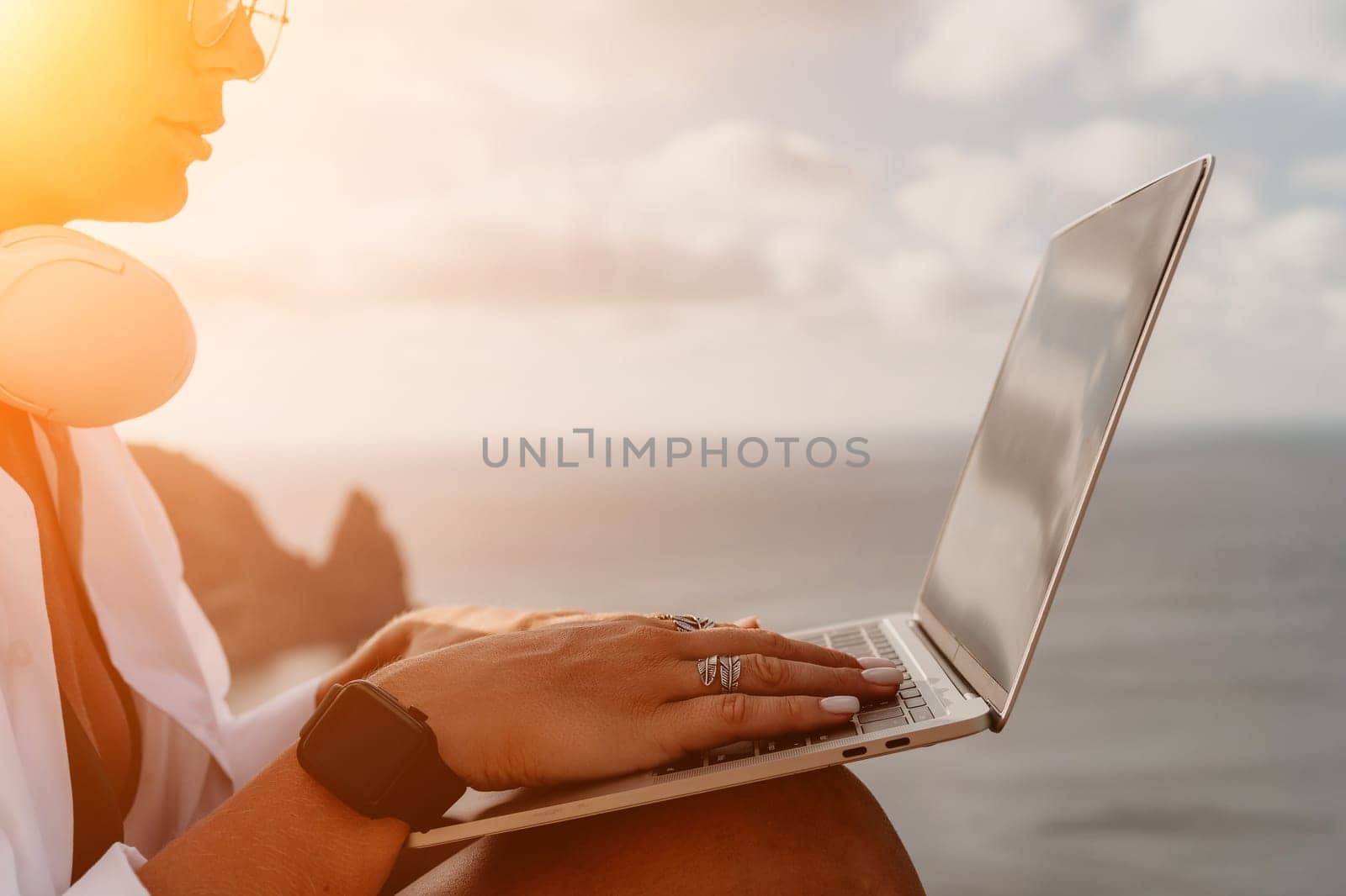 Digital nomad, woman in the hat, a business woman with a laptop sits on the rocks by the sea during sunset, makes a business transaction online from a distance. Freelance, remote work on vacation.