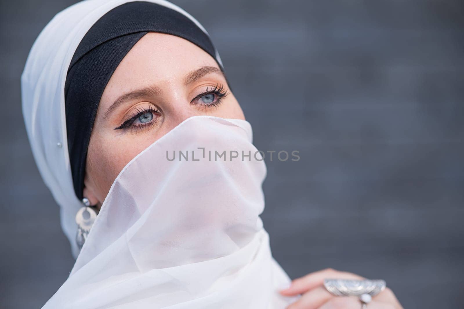 Portrait of a young blue-eyed woman in a hijab against a gray brick wall. Muslim woman with white scarf covering her face
