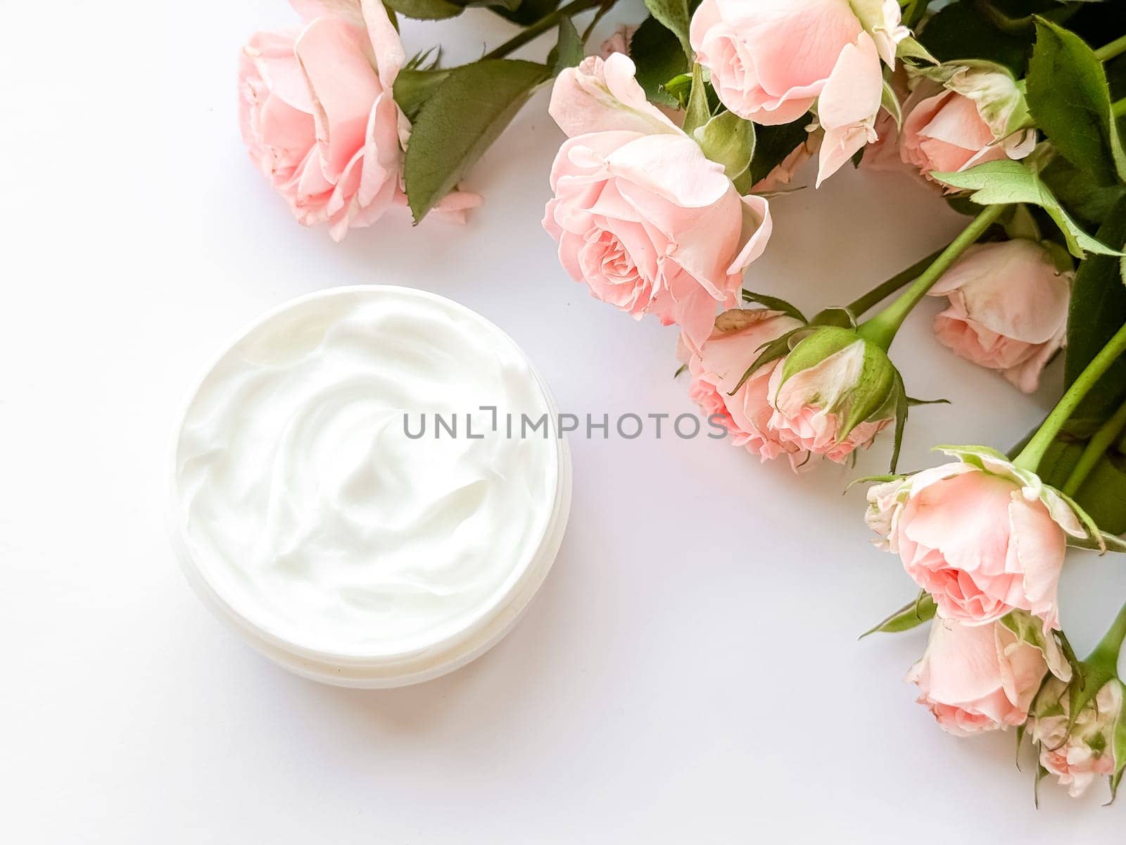 Open jar of white hand, body or face cream next to pink tea roses on white background. Flat lay composition. Skincare and beauty product concept. High quality photo