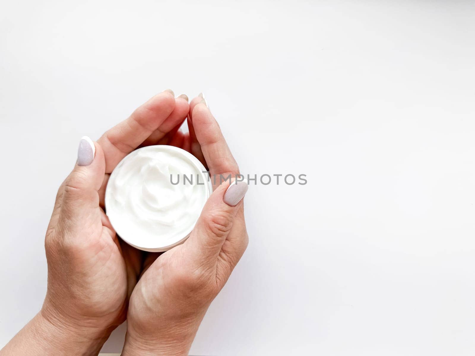 Hands middle age woman hold an open jar of white hand or body cream on white background with copy space. Flat lay composition. Skincare and beauty product concept. Top view. High quality photo