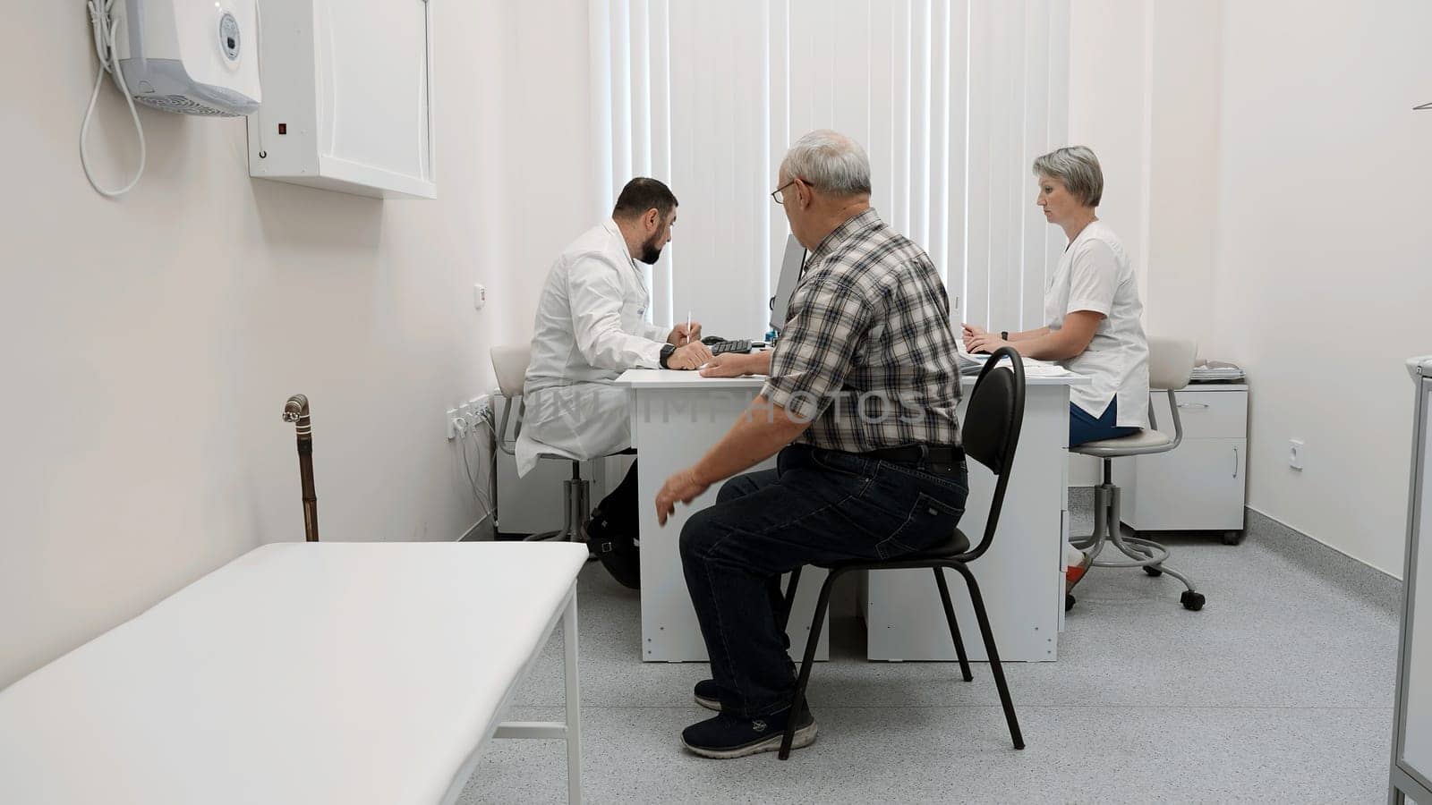 Senior man patient sitting on chair by the table of a doctor and a nurse. Clip. Medical checkup visit at the hospital