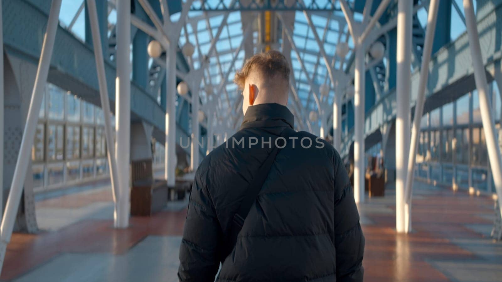 Rear view of man walking in station building. Action. Man walks along sunny corridor of stylish modern building. Beautiful interior of covered bridge of modern city.