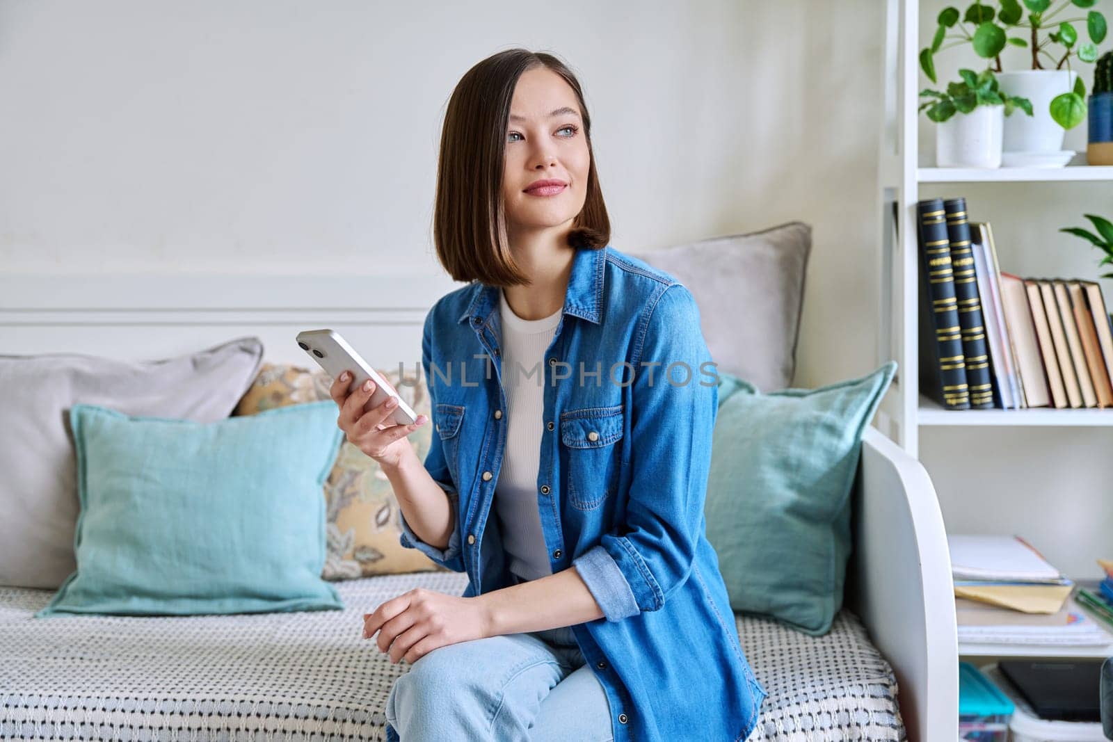 Young woman using smartphone sitting on couch at home. Female texting reading looking, mobile applications technology internet online services for work study leisure communication shopping lifestyle