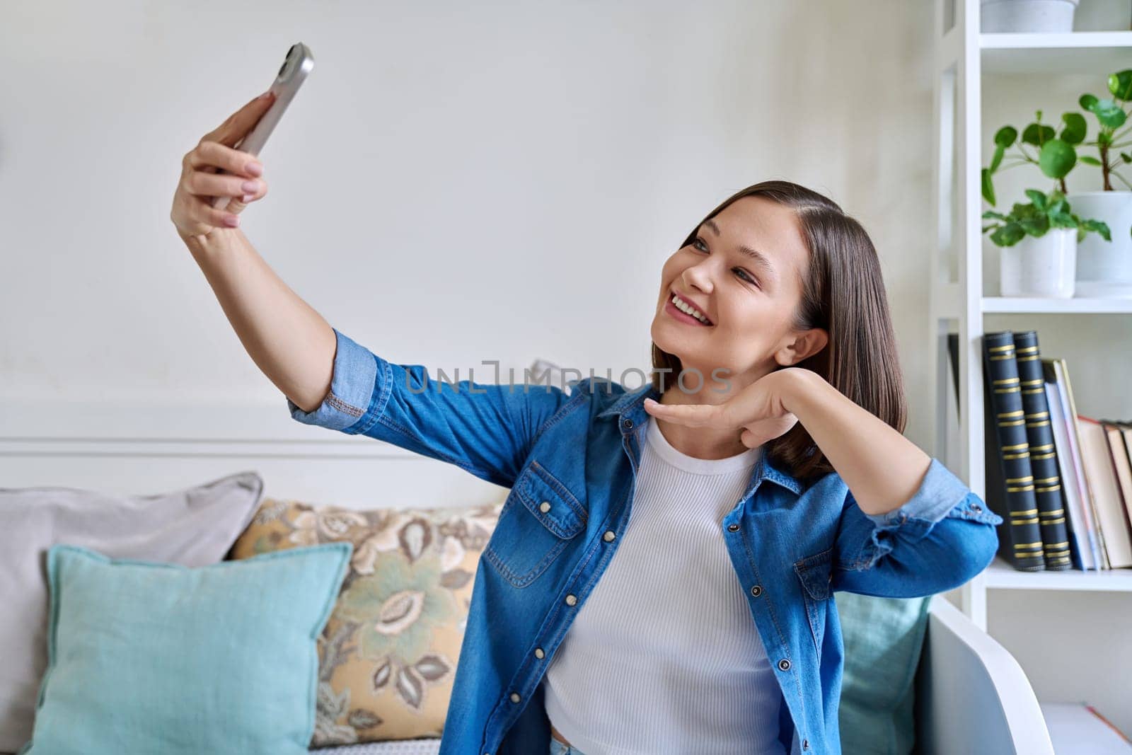 Beautiful happy young woman taking selfie photo on smartphone in home interior. Youth, lifestyle, technology concept