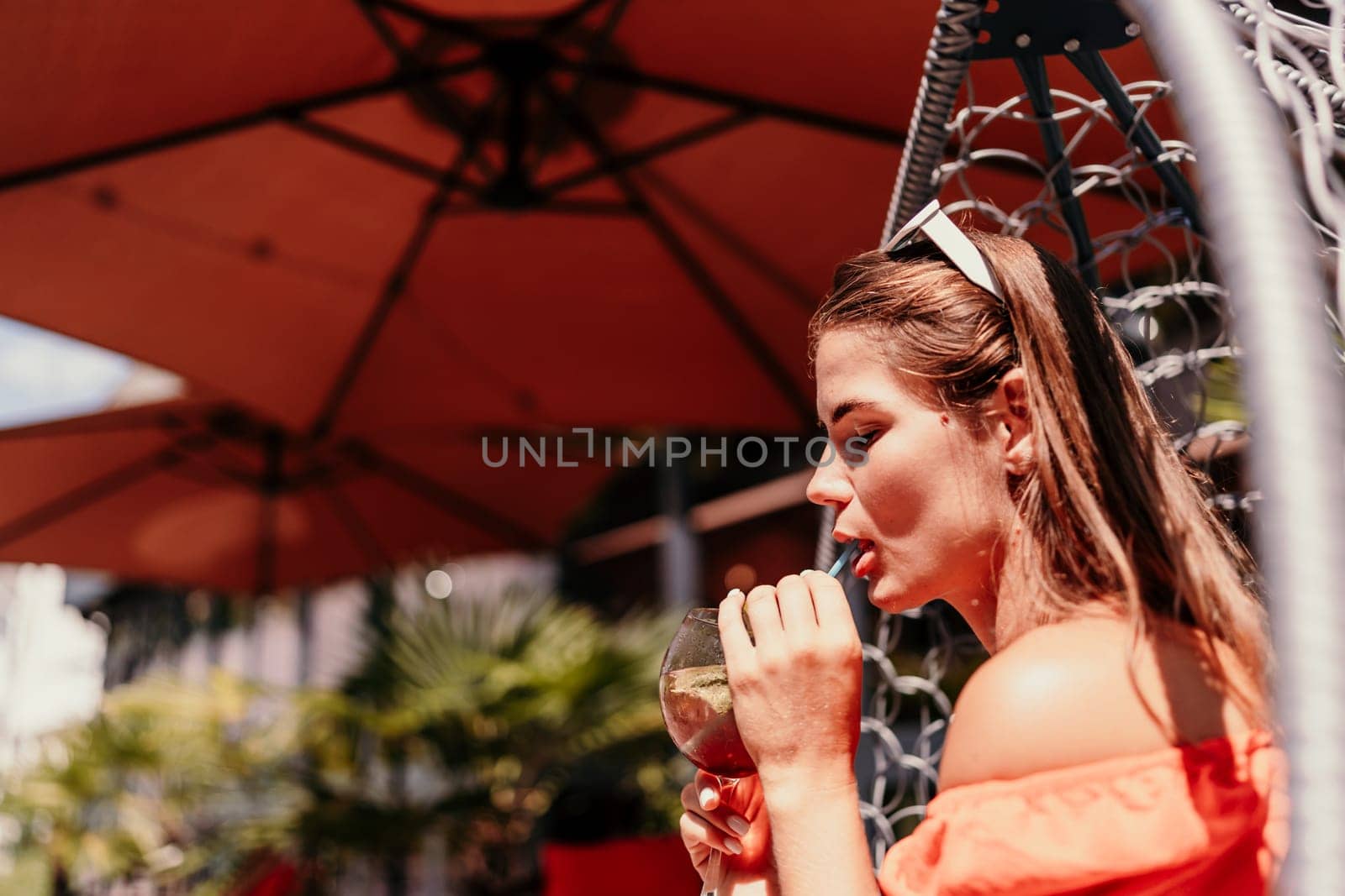 A woman in an orange dress is sitting in a chair with a drink in front of her. She is looking at the camera and she is enjoying her drink. The scene is set in a patio or outdoor area. by Matiunina