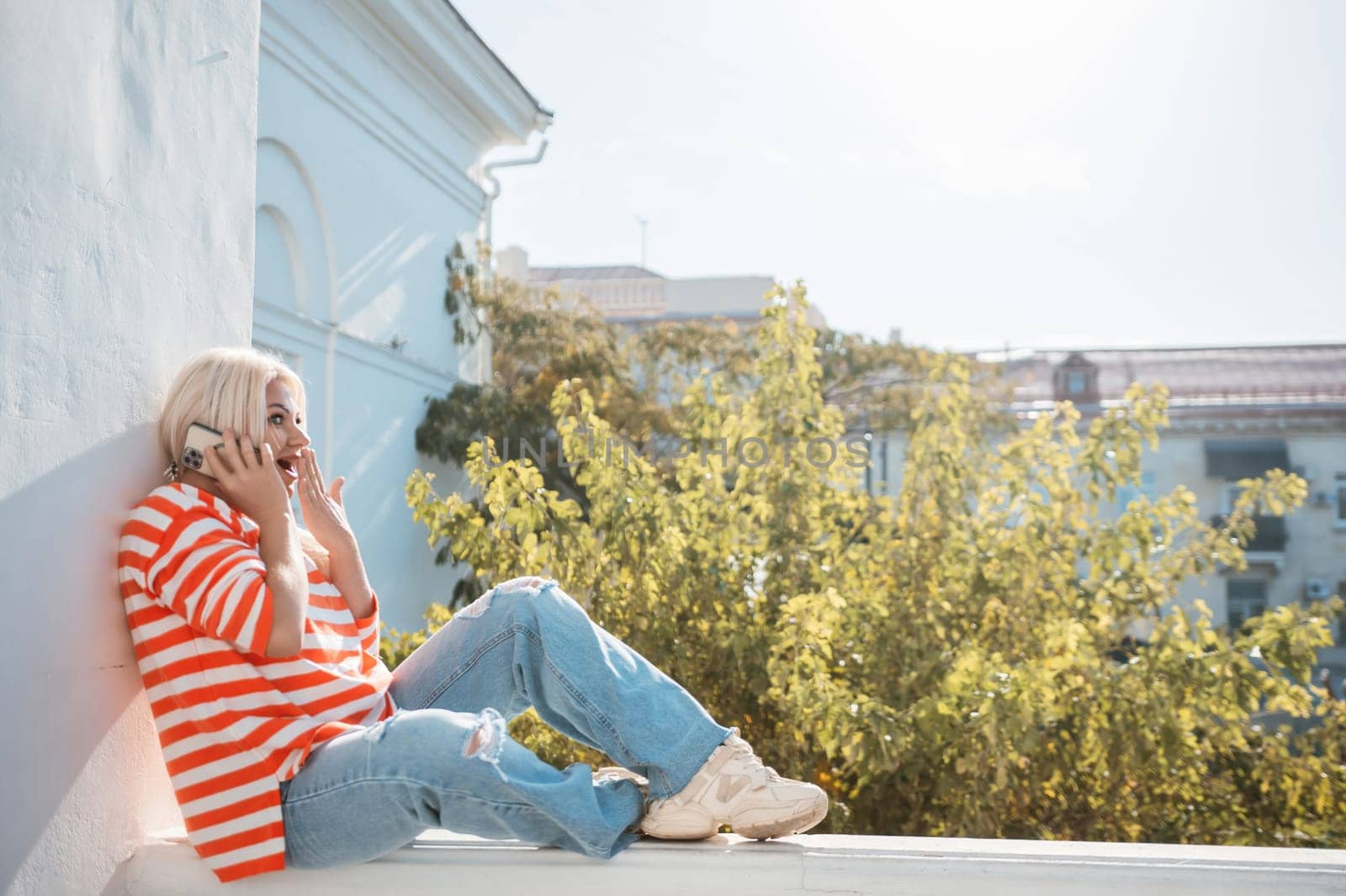 A woman is sitting on a ledge with her phone in her hand. She is wearing a striped shirt and blue jeans. The scene is set in a city with a tree in the background
