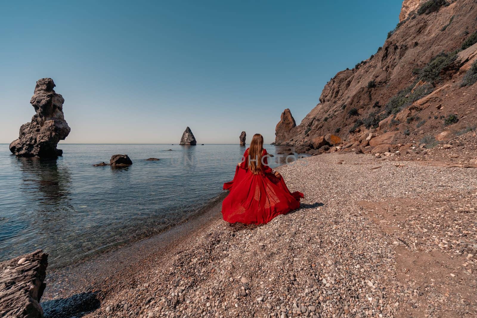 A woman in a red dress stands on a beach with a rocky shoreline in the background. The scene is serene and peaceful, with the woman's red dress contrasting against the natural elements of the beach