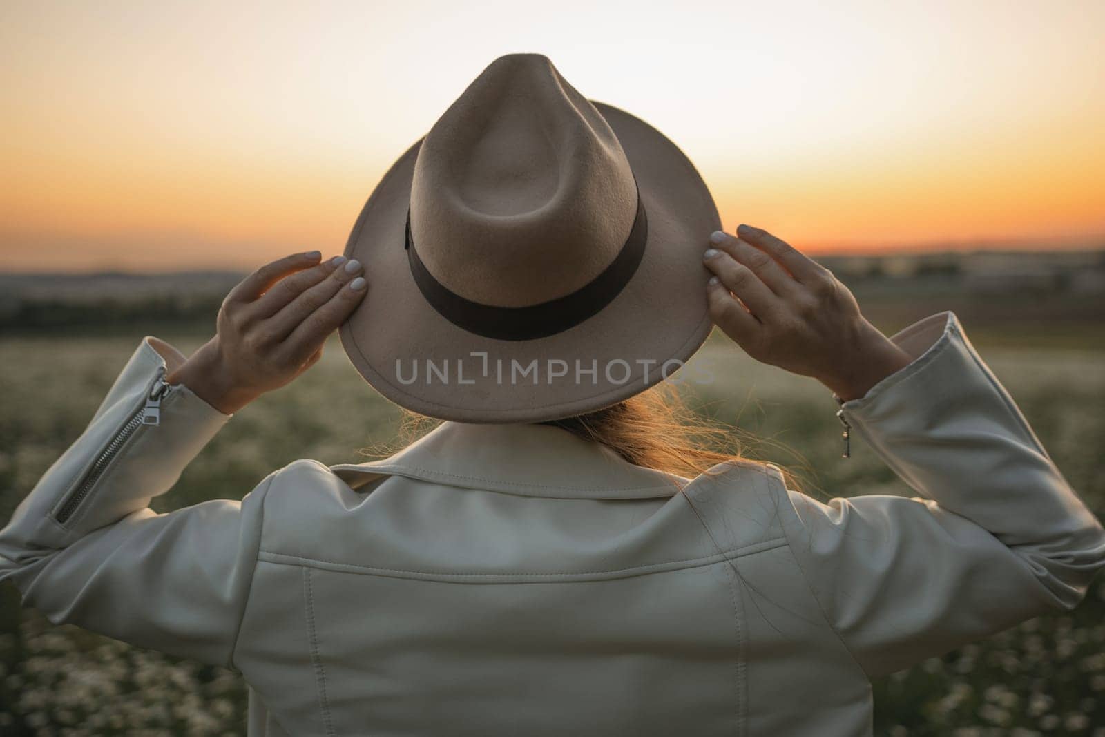 A woman wearing a hat and a white jacket is standing in a field. The woman is looking up at the sky, which is orange and pink. The scene has a peaceful and serene mood. by Matiunina