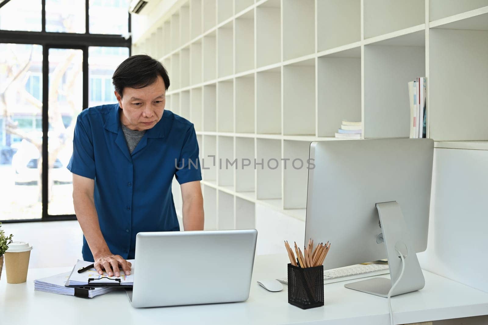 Portrait of senior businessman doing paperwork and using laptop at desk in modern office.