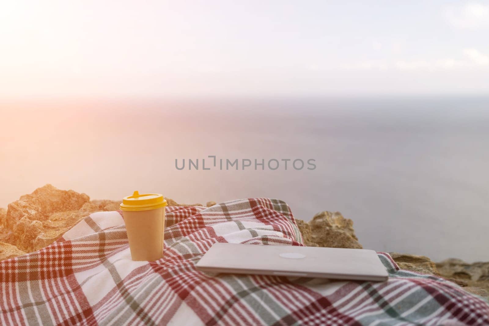 Laptop coffee on blanket with ocean view. Illustrating serene outdoor laptop use. Freelancer enjoying their time outdoors while working or browsing the internet. by Matiunina