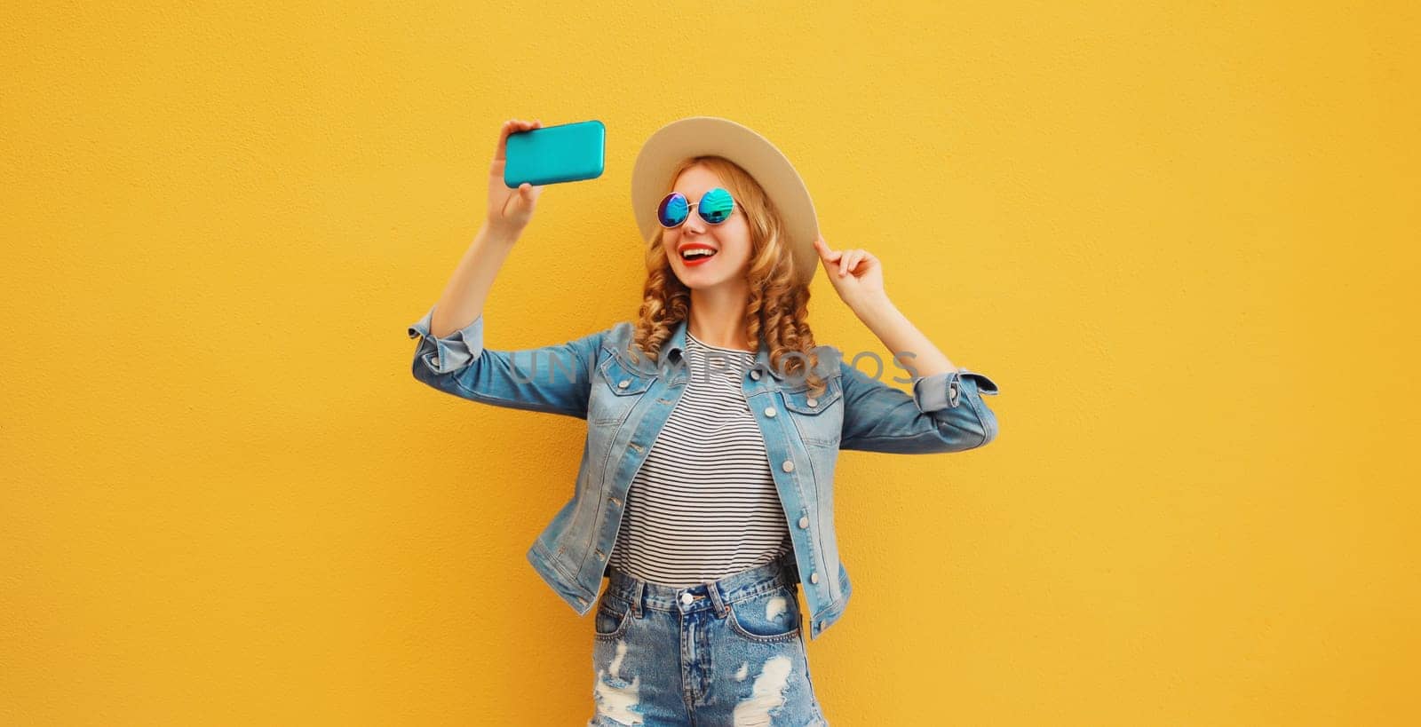 Portrait of happy smiling young woman taking selfie with smartphone wearing summer tourist straw hat, denim clothing on yellow background