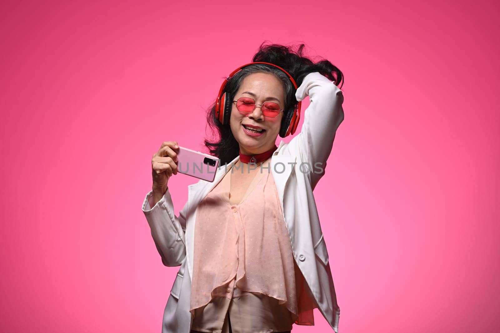 Cheerful elderly woman with headphones enjoying music dancing over pink background.