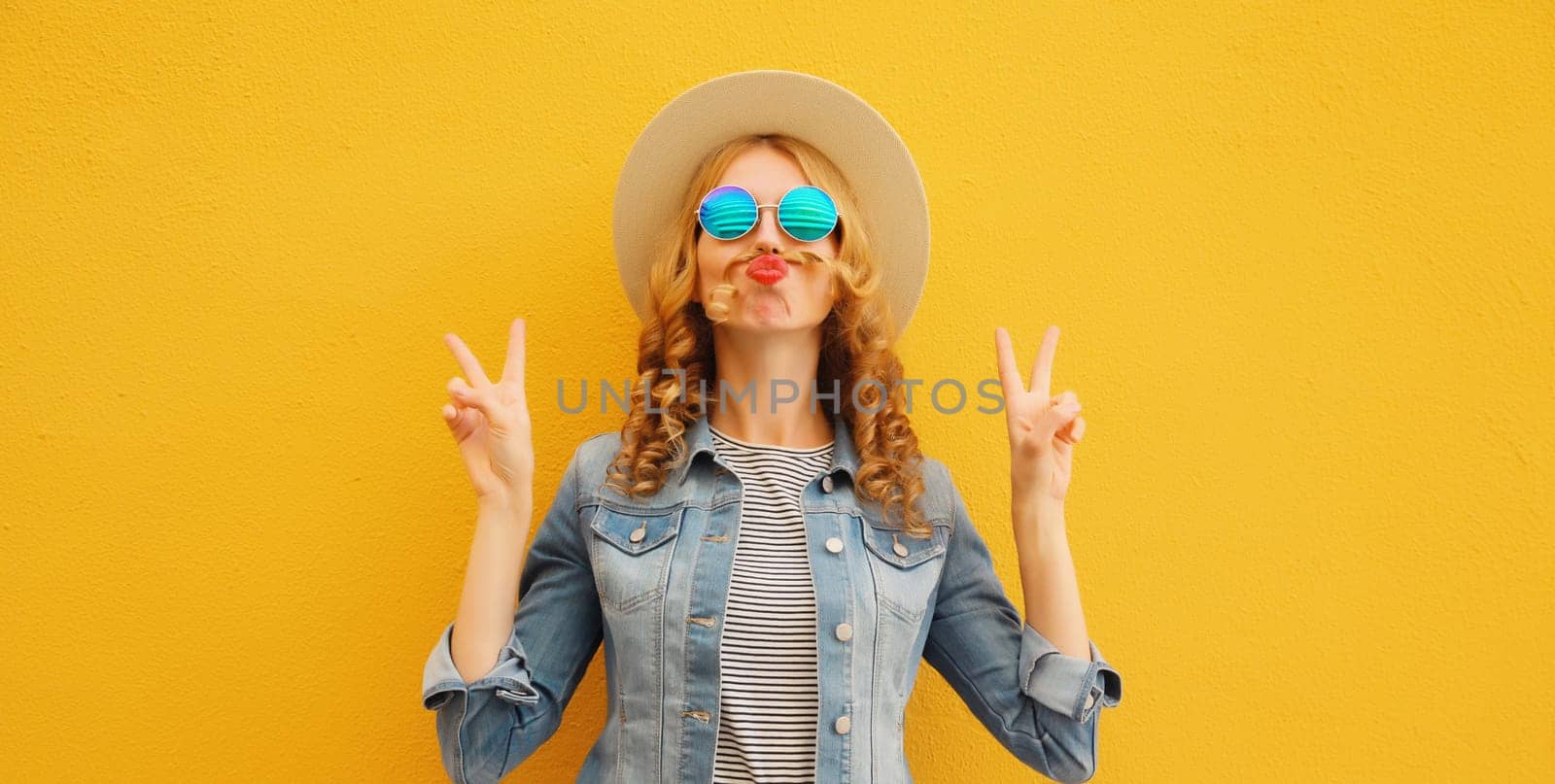 Portrait of stylish funny young woman showing mustache her hair blowing lips with red lipstick wearing summer straw hat on yellow background