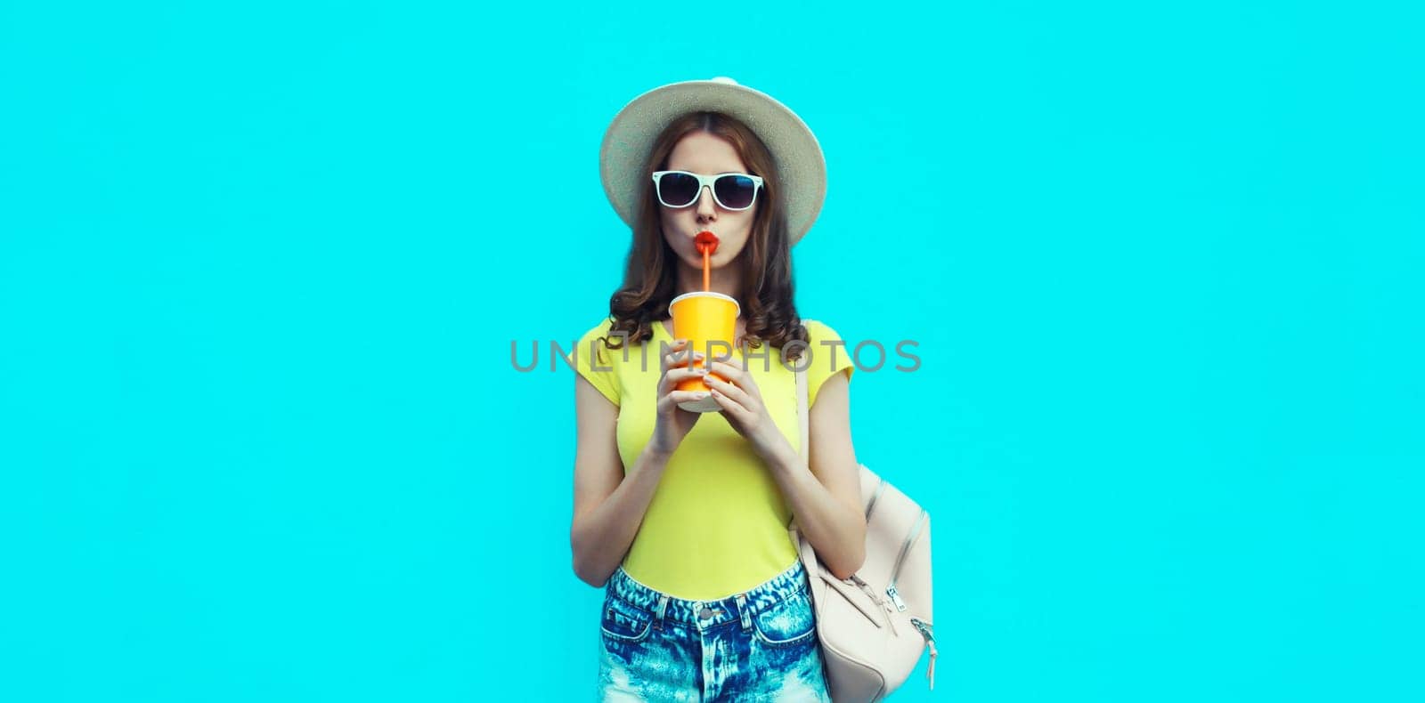 Summer portrait of beautiful caucasian young woman with cup of coffee or fresh juice wearing a straw hat on studio blue background