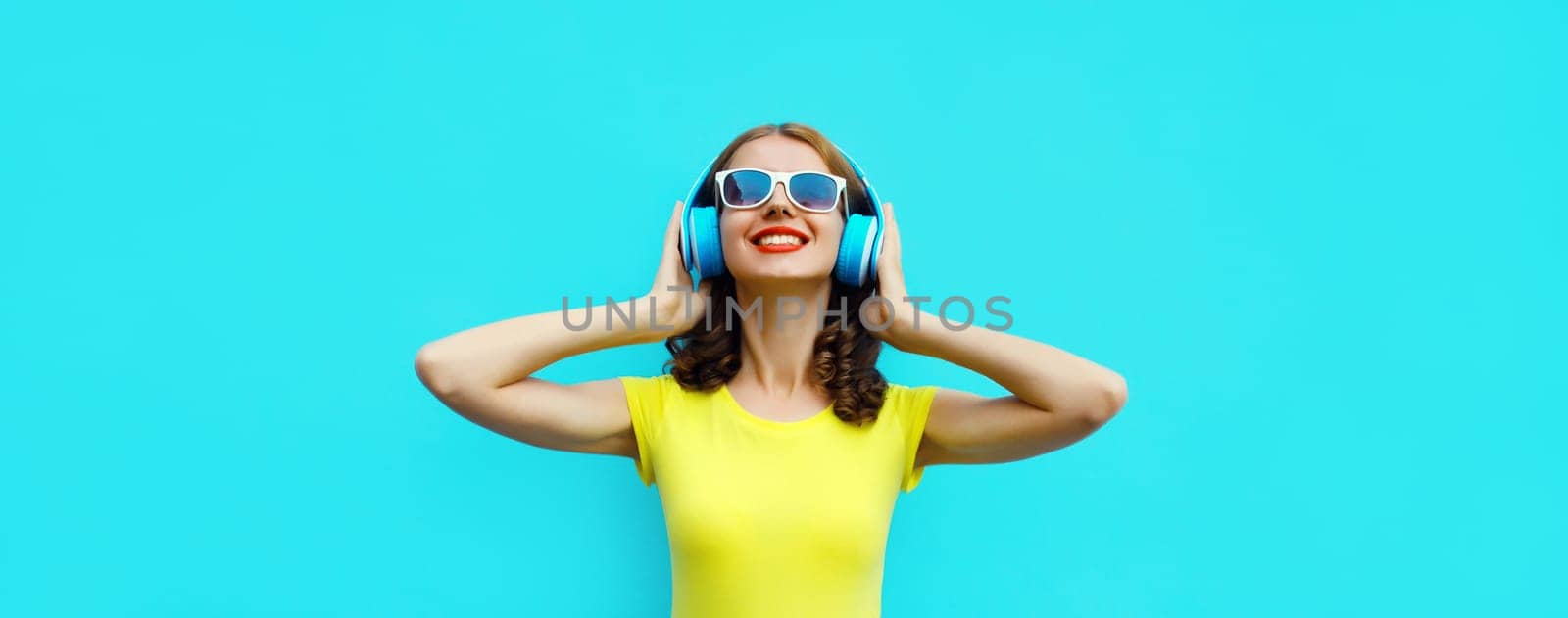 Portrait of happy modern happy young woman listening to music with headphones on blue background