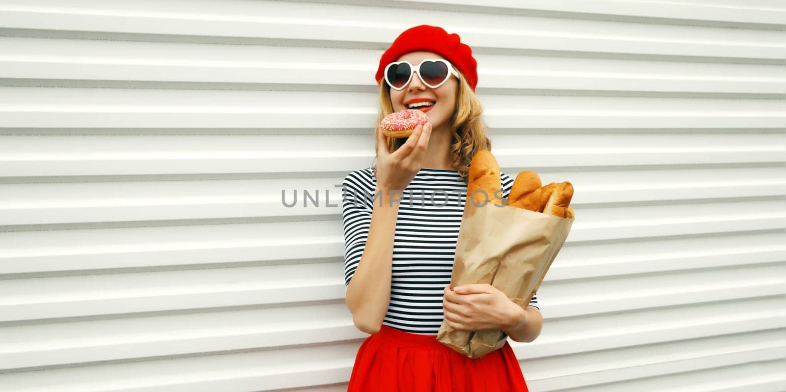 Portrait of happy smiling woman holding grocery shopping paper bag with fresh long white bread baguette eating sweet donut, wear french beret on white background