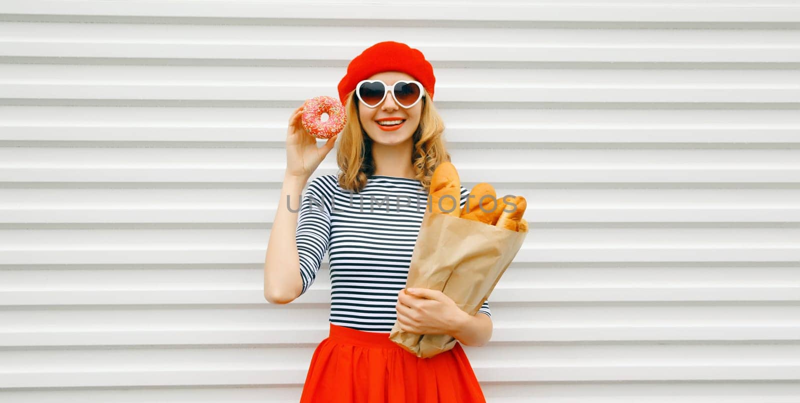 Portrait of happy smiling woman holding grocery shopping paper bag with fresh long white bread baguette eating sweet donut, wear french beret on white background