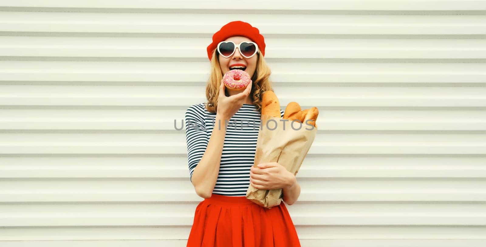 Portrait of happy smiling woman holding grocery shopping paper bag with fresh long white bread baguette eating sweet donut, wear french beret on white background