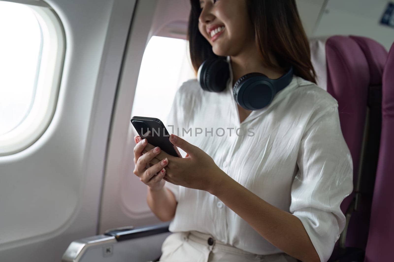 Beautiful Asian in aeroplane. working, travel, business concept Traveler on Plane Using Smartphone with Headphones, Smiling and Happy.