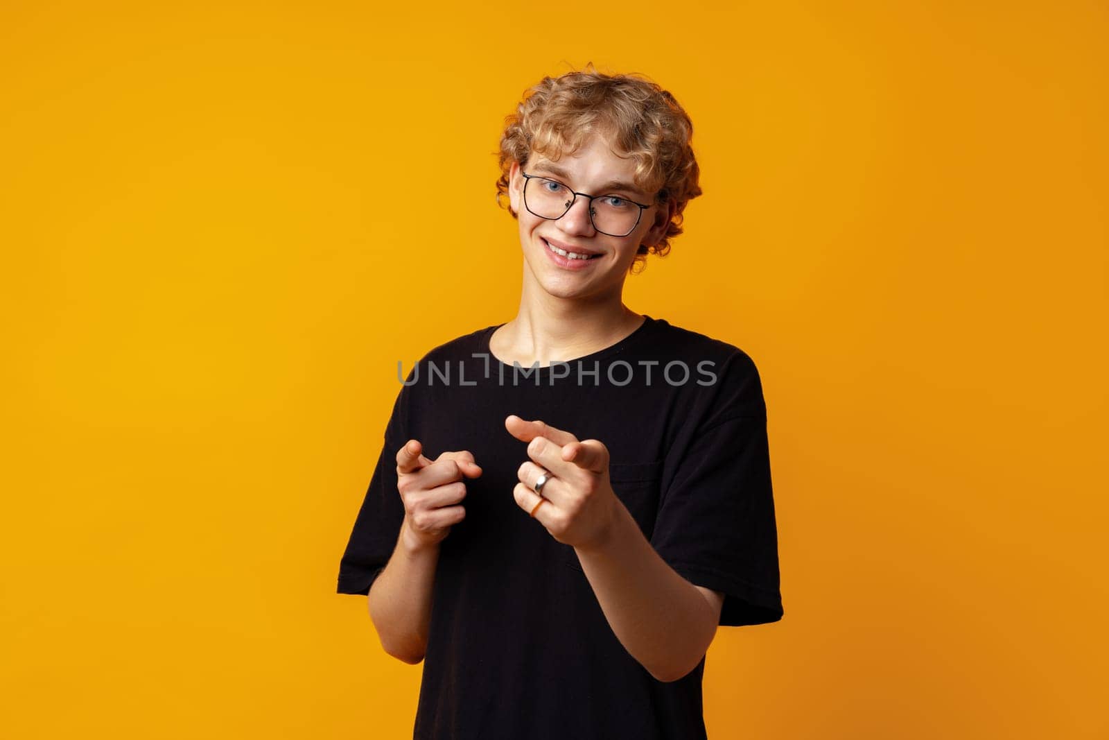 Young curly-haired man in glasses pointing to camera against yellow background close up
