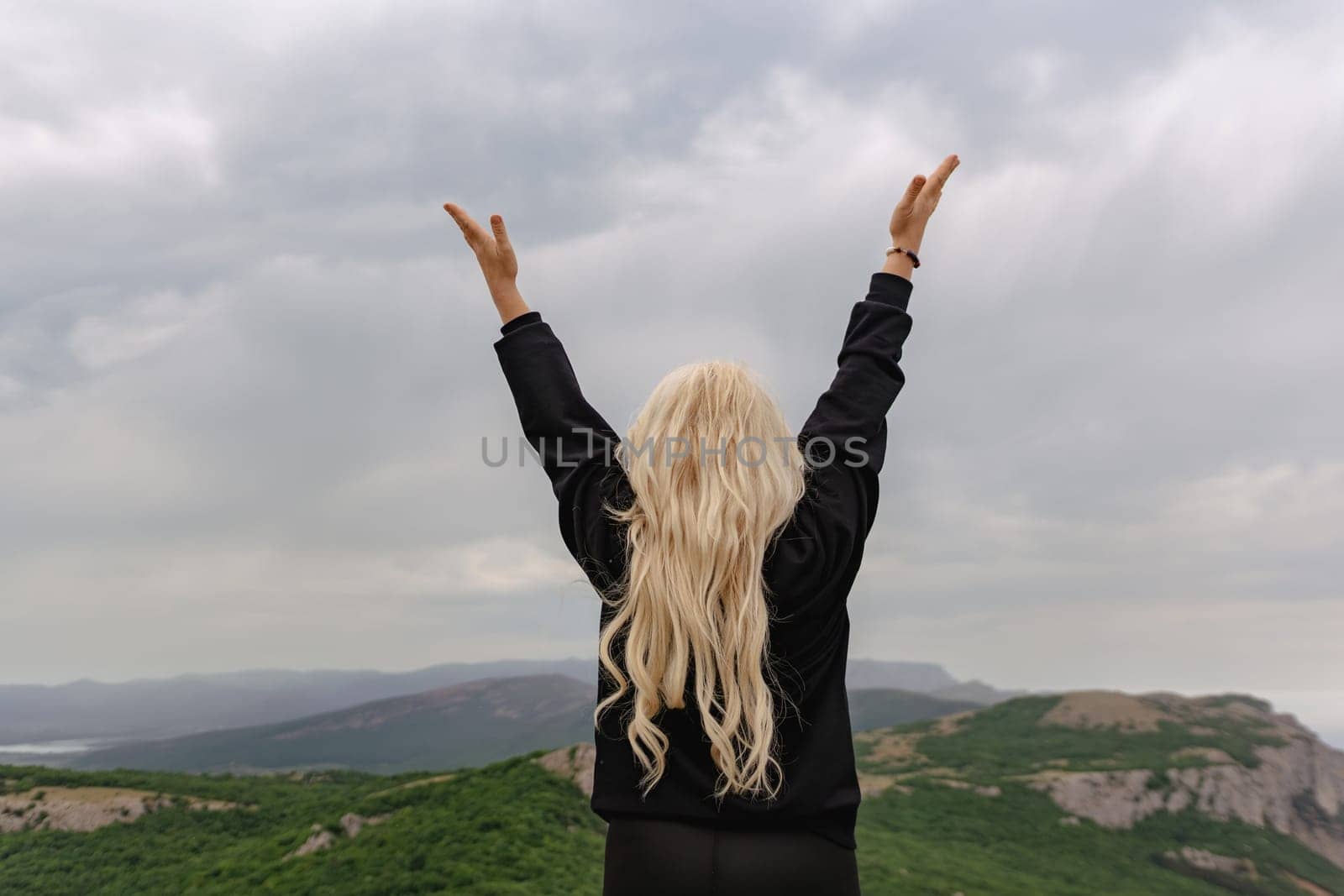 A woman with blonde hair is standing on a mountain and raising her hands in the air. The sky is cloudy, and the mountains are in the background. The woman is happy and joyful