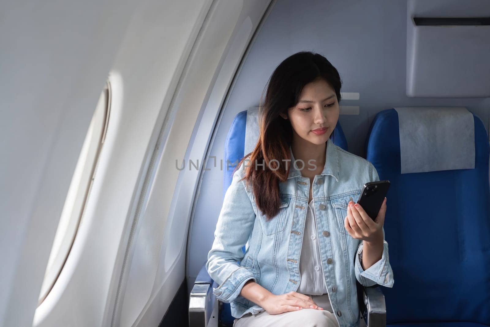 Asian woman using smartphone while sitting in airplane seat. Concept of air travel, technology, and connectivity by wichayada