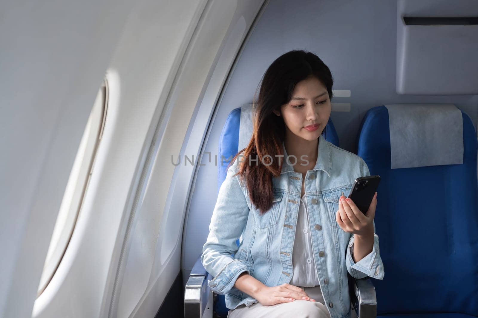 Asian woman using smartphone while sitting in airplane seat. Concept of air travel, technology, and connectivity.