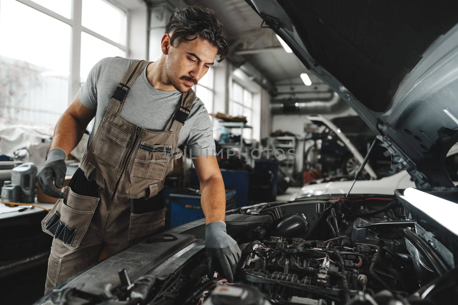 Young male mechanic examining engine under hood of car at the repair garage close up