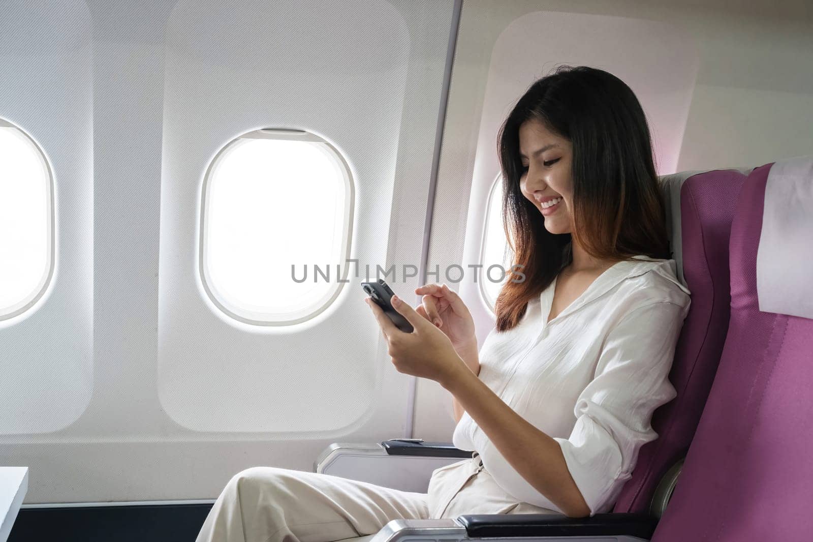 Asian woman using smartphone on airplane seat during flight. Concept of air travel, technology, and connectivity by wichayada